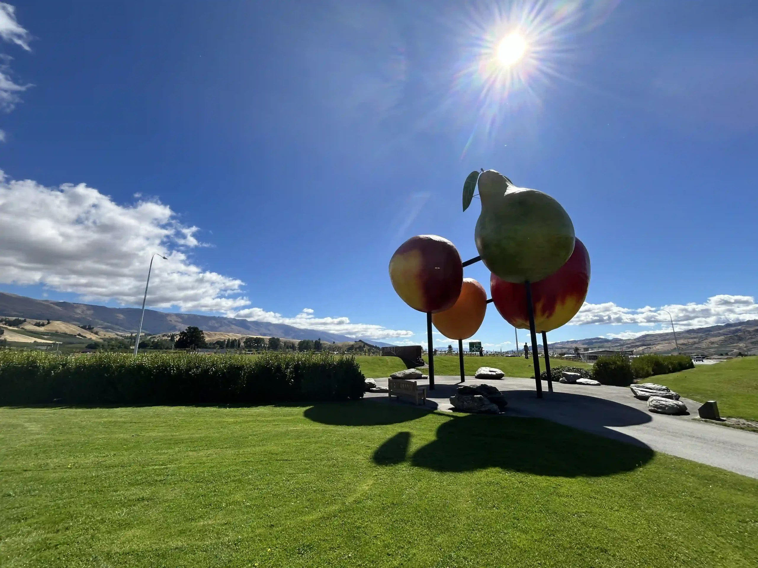 Cromwell South Island giant fruit sculpture at the main roundabout — oversized pear, nectarine and apple on metal poles against blue sky and Central Otago hills
