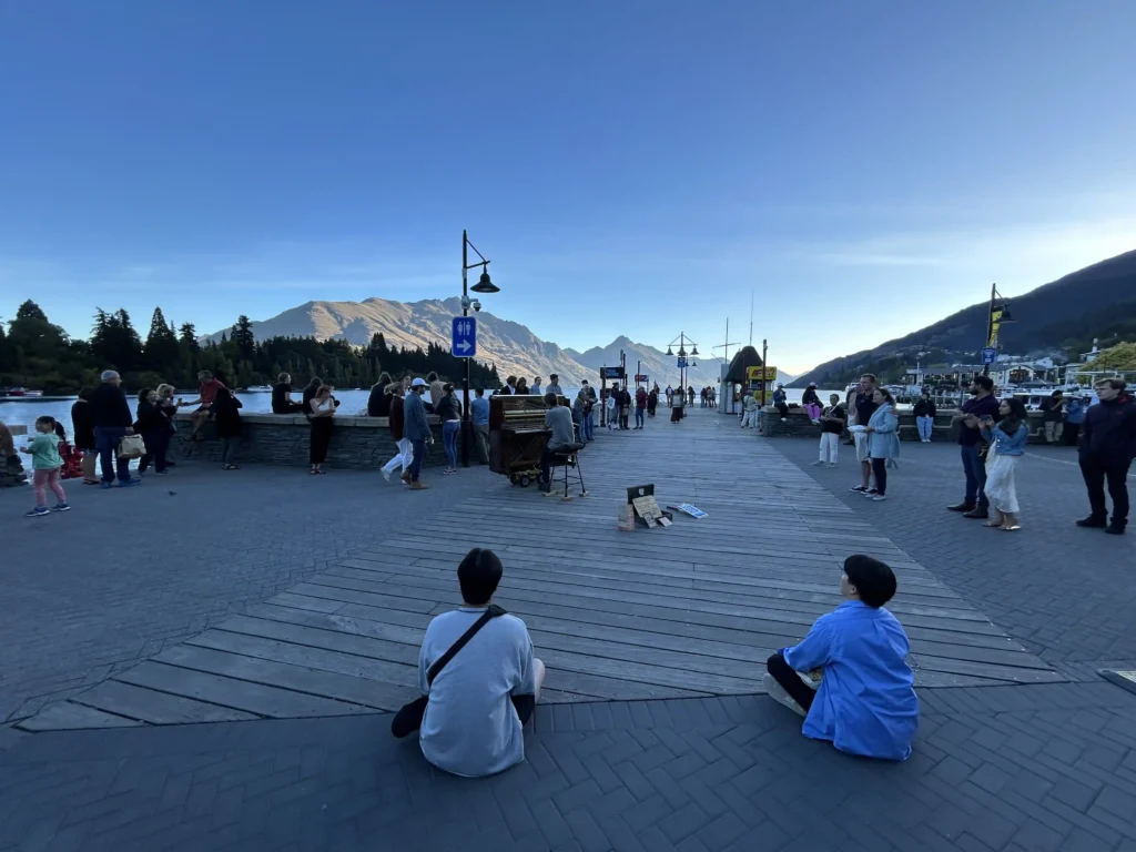 Piano busker performing at Queenstown lakefront pier at sunset — crowd gathered around, two children sitting on the ground listening